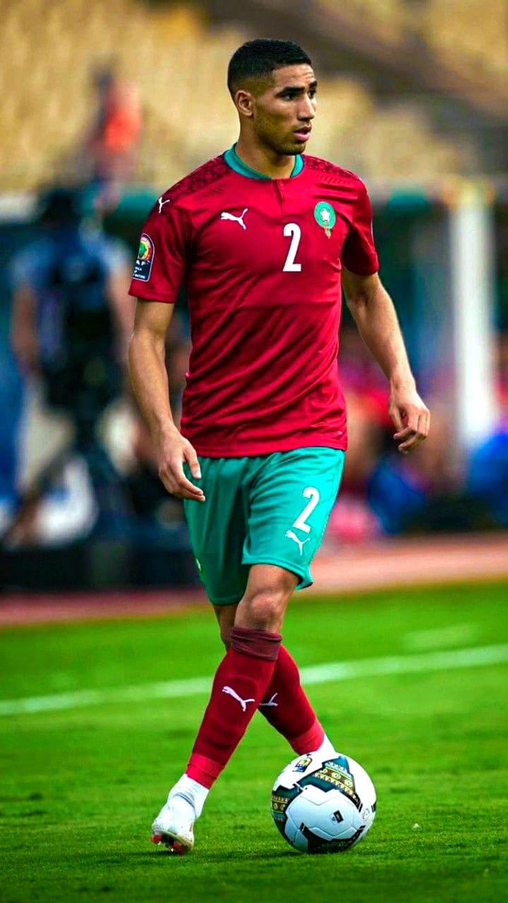 A soccer player in a red and green Morocco uniform with the number 2 controls the ball on a field during a match, as fans around the world tune in via Croatia IPTV to catch every moment of the action.
