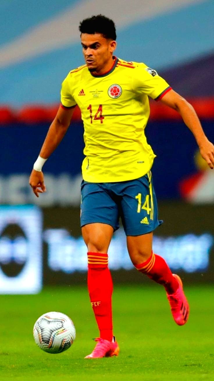 A soccer player in a yellow jersey with the number 14 controls the ball on a field. Wearing blue shorts, red socks, and pink shoes, he appears focused during a match broadcasted via Croatia IPTV.